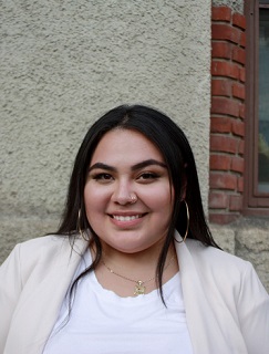 young woman smiling while posing in front of building. She has light medium skin tone, jewelry, white shirt and blazer.
